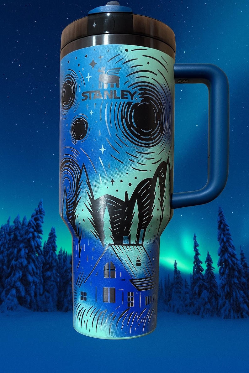 Puede incluir: Un termo de viaje Stanley azul y verde azulado con asa, con un dise&ntilde;o de paisaje invernal. El dise&ntilde;o incluye una casa, &aacute;rboles y patrones arremolinados, con el logotipo de Stanley. El termo tiene una tapa negra.