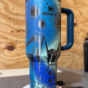Puede incluir: Un termo Stanley con un dise&ntilde;o de noche estrellada azul y negro. El termo tiene un asa y tapa azul oscuro. El dise&ntilde;o incluye una casa, &aacute;rboles y estrellas. El logotipo de Stanley es visible.