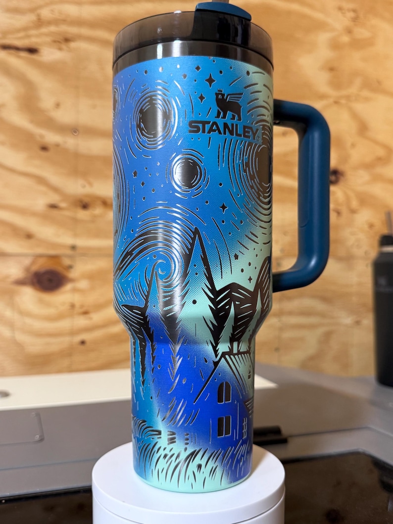 Puede incluir: Un termo Stanley con un dise&ntilde;o de noche estrellada azul y negro. El termo tiene un asa y tapa azul oscuro. El dise&ntilde;o incluye una casa, &aacute;rboles y estrellas. El logotipo de Stanley es visible.