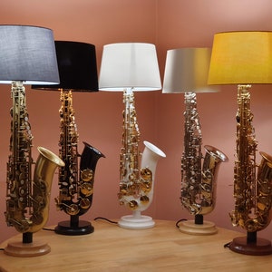 May include: Five table lamps shaped like saxophones with different colored lampshades. The lampshades are gray, black, white, white, and yellow. The saxophones are gold, silver, and black.