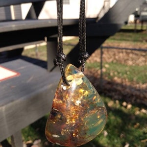 May include: A teardrop-shaped pendant necklace with a green, brown, and gold amber stone. The stone is translucent and has inclusions of air bubbles and other organic matter. The necklace is made with a black cord.