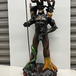 May include: A three-headed horned figure statue with a black torso, one yellow bird-like leg, and one human-like leg. The figure holds a tall trident, features a gold-colored sulfur symbol on the chest, and stands on a base with flames and skulls.