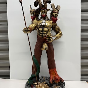 Könnte beinhalten: Eine dreiköpfige gehörnte Figur mit goldener Haut, rotem Bart und muskulösem Oberkörper, die eine silberne Leviathan-Kreuz-Halskette trägt. Die Figur hält einen hohen Dreizack, steht auf einem strukturierten runden Sockel und hat eine grüne Schlange, die sich um ein Bein windet.