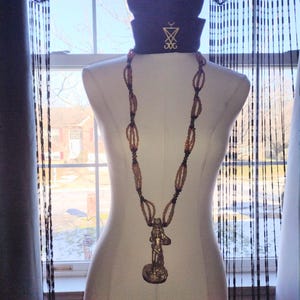 May include: A navy blue beanie with a gold emblem and a long beaded necklace with a pendant of a figure. The necklace is draped on a white mannequin. The background includes a window and beaded curtains.