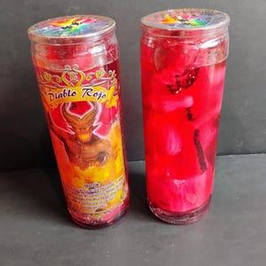 Puede incluir: Dos velas de vidrio rojo con cera roja y decoraciones blancas y rojas. La vela de la izquierda tiene una etiqueta que dice "Diablo Rojo" y una imagen de un diablo rojo. La vela de la derecha tiene un palo negro en el centro.