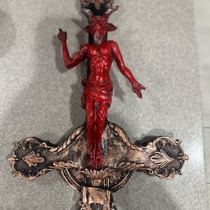 Puede incluir: Una figura roja con cuernos y barba está representada en una cruz negra y dorada. La figura está en una pose crucificada con los brazos extendidos. La cruz tiene un diseño decorativo con detalles ornamentados.