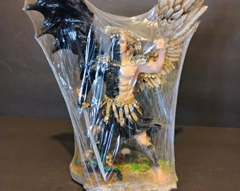 Puede incluir: Figura de un ángel guerrero con una espada, con un ala negra a la izquierda y un ala dorada a la derecha. El ángel está representado con un físico musculoso, vistiendo una armadura negra y detalles dorados. La figura está sobre una base de piedra.
