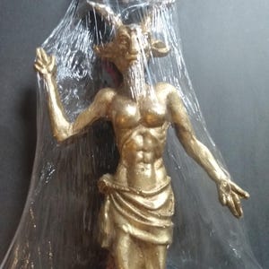 Op de afbeelding: Een gouden beeld van een gehoornde figuur met een gespierd lichaam. Het beeld is verpakt in doorzichtig plastic en lijkt een decoratief item te zijn.