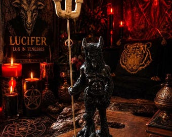 May include: A black horned figure statue standing on a rocky base, holding a tall, thin gold-colored rod topped with a three-pronged cream-colored trident. The figure has a textured, dark body with prominent horns and stands in a vertical, upright pose.