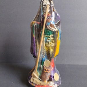 Peut inclure: Une figurine colorée de Santa Muerte, la sainte populaire mexicaine de la mort, tenant une faux et un bâton. La figurine est en résine et est peinte dans des couleurs vives, notamment le violet, le vert, le jaune et l'or. La figurine porte une longue robe flottante et a un visage de crâne.
