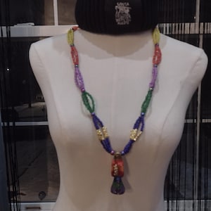 May include: A beaded necklace with a pendant is displayed on a mannequin. The necklace features colorful beads in red, yellow, green, purple, and blue. The pendant is a mix of red, orange, and purple. A black beanie with text is worn on the mannequin.