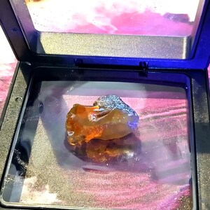 Op de afbeelding: Een doorschijnende, onregelmatig gevormde barnsteenkleurige edelsteen rust in een zwarte vierkante vitrine. De steen vertoont interne gouden en oranje tinten die licht reflecteren op het glanzende oppervlak van de bodem van de houder.