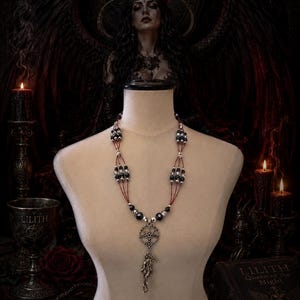May include: A multi-strand necklace featuring dark faceted beads and silver-toned spacers, centred with a circular pentagram pendant and a dangling figure charm. The necklace is displayed on a neutral-toned mannequin against a dark background.