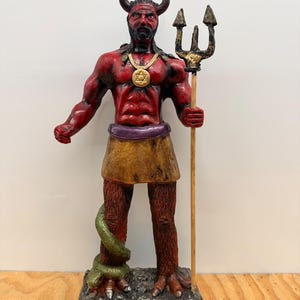 Puede incluir: Una estatua de figura con cuernos y piel roja sobre una base con calaveras. La figura lleva un collar dorado con una Estrella de David, un cinturón morado y una falda color canela. Una serpiente verde se enrosca en una pierna y sostiene un tridente.