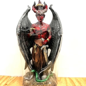 Könnte beinhalten: Eine rot häutige, geflügelte Figurenstatue mit Hörnern, die einen goldenen Gürtel trägt und ein silbernes Schwert hält. Die Figur hat große schwarze Flügel, eine grüne Schlange zu ihren Füßen und einen Sockel mit der Inschrift LUZ, alles in Plastik.