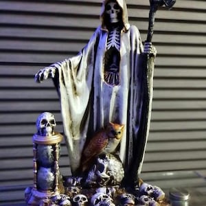 May include: A detailed figurine of the Grim Reaper, clad in a white hooded robe, holding a scythe. The base features skulls, an owl, and an hourglass. The statue is painted in shades of white, gray, brown, and gold, creating a spooky aesthetic.