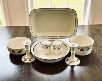 May include: A cream-colored dinnerware set with a black trim and floral accents. The set includes a rectangular baking dish, two bowls, a serving plate, salt and pepper shakers, and two candle holders.