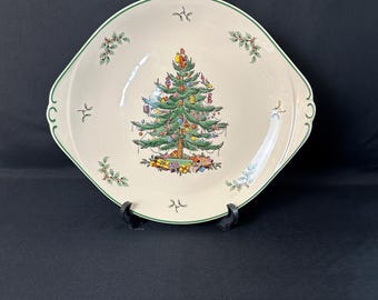 May include: Cream-colored, square-shaped serving dish with a green rim and handles. The center features a decorated Christmas tree with ornaments and presents. Holly and berry sprigs are placed around the rim, creating a festive holiday theme.