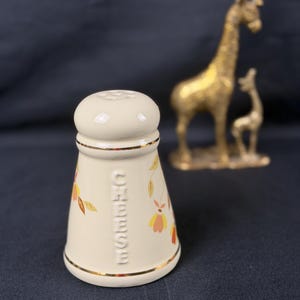 Puede incluir: Salero crema con un diseño floral en naranja y amarillo, y la palabra "Chinese" en relieve en el lateral. El salero tiene un borde dorado y una tapa a juego. En el fondo, hay dos figuras de jirafas doradas.