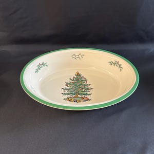 May include: Oval serving dish with a Christmas tree design. The white ceramic dish has a green border and a detailed illustration of a decorated Christmas tree with presents underneath. Small sprigs of holly are also featured.