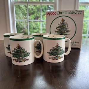 May include: Four white Spode Christmas Tree mugs with green trim and handles, each featuring a decorated Christmas tree design. A matching box is visible in the background, with the words "Spode Christmas Tree" and a tree illustration.
