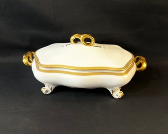 May include: White porcelain serving dish with a gold rim and ornate gold handles. The lid has a gold knob and a delicate floral design.