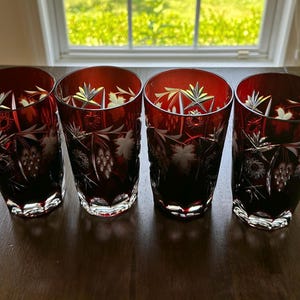 May include: Four ruby red crystal drinking glasses with etched floral and grape vine designs. The glasses have a faceted base and a tall, straight shape. The glasses are arranged on a dark wooden surface.