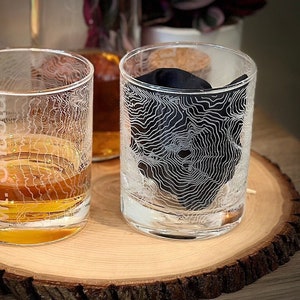 May include: Two clear whiskey glasses with etched topographical map designs. One glass is filled with amber liquid, the other holds a black cloth. The glass on the left has the text "Mt. Shasta". Both glasses sit on a wooden slab.