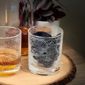May include: Two clear glass tumblers with a topographic map design etched on the side. The tumblers are sitting on a wooden coaster.