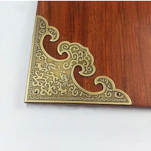 May include: A gold-colored metal corner bracket with an intricate design featuring swirling patterns and a geometric border. The bracket is designed to be used as a decorative accent for furniture or other wooden objects.