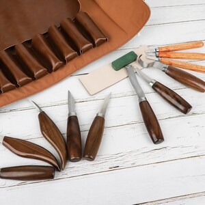 Pu&ograve; includere: Un set di strumenti per intagliare il legno con manici in legno e lame in metallo, insieme a un rotolo di pelle per la conservazione. Gli strumenti sono disposti su una superficie di legno bianco con una pietra per affilare verde.