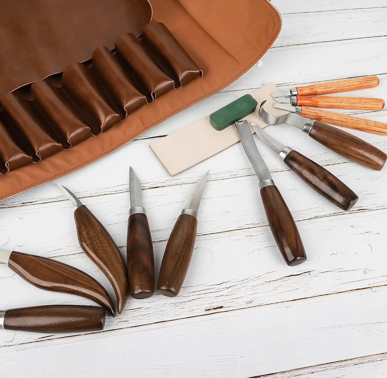 Pu&ograve; includere: Un set di strumenti per intagliare il legno con manici in legno e lame in metallo, insieme a un rotolo di pelle per la conservazione. Gli strumenti sono disposti su una superficie di legno bianco con una pietra per affilare verde.