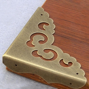 May include: Antique brass corner bracket with a decorative, ornate design. The bracket has a scalloped edge and a swirl pattern.