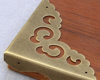 May include: Antique brass corner bracket with a decorative, ornate design. The bracket has a scalloped edge and a swirl pattern.