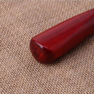 Puede incluir: Primer plano del mango de un bate de b&eacute;isbol de madera. El mango es de un color rojo intenso y rico con vetas de madera visibles. El extremo del mango es redondeado y pulido, reflejando la luz. El fondo es una tela texturizada de color beige.