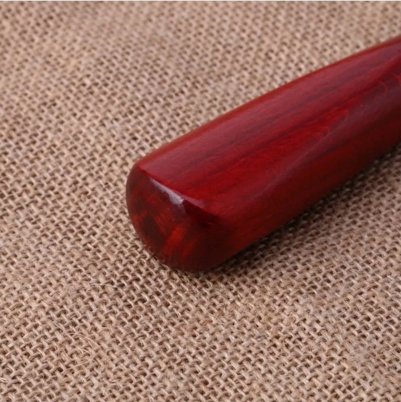Puede incluir: Primer plano del mango de un bate de b&eacute;isbol de madera. El mango es de un color rojo intenso y rico con vetas de madera visibles. El extremo del mango es redondeado y pulido, reflejando la luz. El fondo es una tela texturizada de color beige.