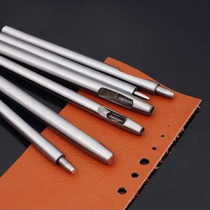 May include: Set of six silver leather hole punch tools of varying sizes. The tools are arranged on a brown leather surface with several holes punched in it.