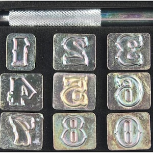 May include: A set of ten metal letter stamps in a black case with a silver handle. The stamps are numbered 1 through 0 and have a decorative, vintage-style font.