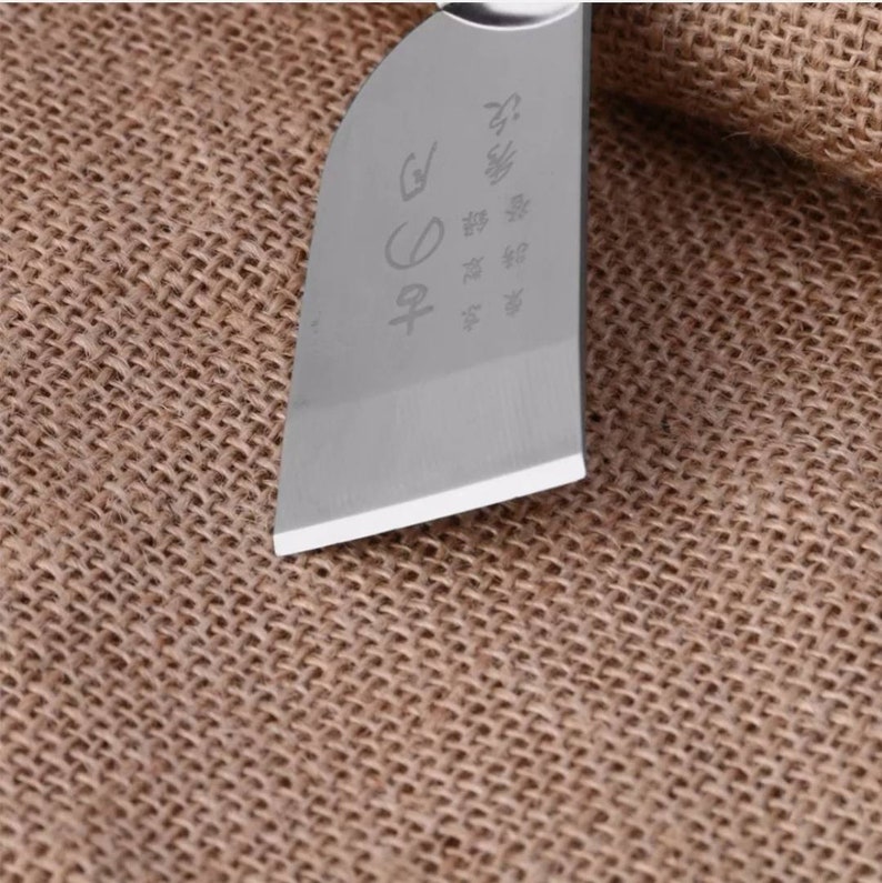 Puede incluir: Primer plano de una hoja de cuchillo de acero inoxidable con caracteres japoneses grabados. La hoja tiene un filo afilado y un acabado pulido. El cuchillo est&aacute; sobre un fondo tejido marr&oacute;n.