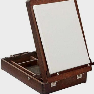 May include: A wooden artist's easel with a white canvas. The easel is dark brown and has a hinged lid that opens to reveal storage compartments. The canvas is held in place by a wooden frame and metal clips. The easel is designed for painting and art projects.
