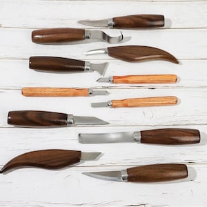 Pu&ograve; includere: Un set di nove strumenti per intagliare il legno con manici in legno. Gli strumenti hanno diverse forme e dimensioni delle lame e sono disposti su una superficie in legno bianco.