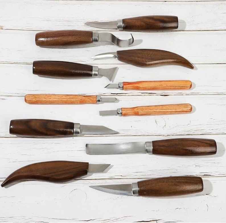 Pu&ograve; includere: Un set di nove strumenti per intagliare il legno con manici in legno. Gli strumenti hanno diverse forme e dimensioni delle lame e sono disposti su una superficie in legno bianco.