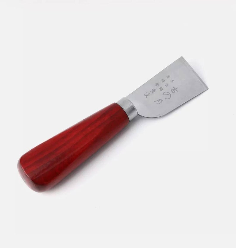 Puede incluir: Cuchillo para artesan&iacute;a en cuero con una hoja de acero pulido y un mango de madera rojo oscuro redondeado. La hoja tiene caracteres japoneses grabados. El mango mide unos 10 cm.