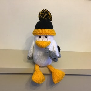 May include: A white knitted seagull wearing a black and yellow knitted hat with a yellow pom pom. The seagull is holding a grey knitted fish in its beak and is sitting on a shelf.