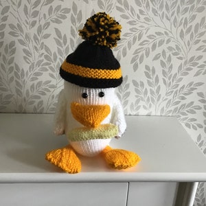 May include: A white knitted penguin wearing a black and yellow knitted hat with a black and yellow pom pom. The penguin is holding a green and white knitted object in its beak.