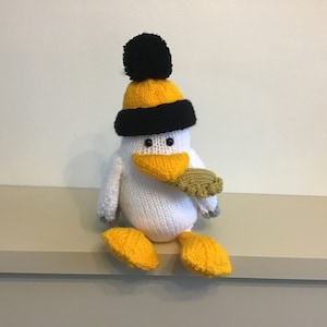 May include: A white knitted toy bird wearing a yellow and black knit hat with a black pom-pom. The bird has a yellow beak and yellow feet.