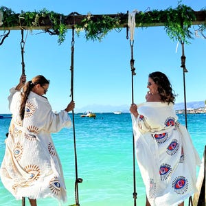 Puede incluir: Dos personas en columpios en el mar, vistiendo túnicas blancas con diseños de ojos. Los columpios están sujetos a una estructura de madera con las palabras "PODA KSAMIL". El agua es de color turquesa.