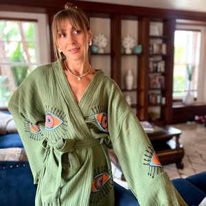 May include: Olive green robe with an eye design. The robe features a wrap-around style with a matching belt and fringe detailing on the sleeves. The eye design is in orange, pink, and black. The robe is made of a lightweight fabric.