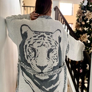 May include: A light-colored kimono-style robe with a large tiger face design on the back in shades of gray. The robe has a matching belt and decorative trim along the edges. The sleeves are wide and flowing.
