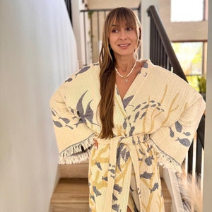 May include: A woman wearing a white robe with a blue and yellow fish print. The robe has a sash tie and fringe trim. She is standing on a staircase.